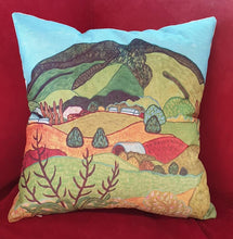 Load image into Gallery viewer, A square decorative throw pillow featuring a colorful landscape design with green hills, red fields, and a light blue sky, displayed on a red velvet chair.