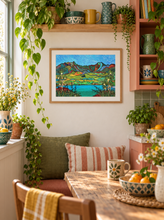 Load image into Gallery viewer, A colorful framed landscape art print hangs on a white wall in a cozy, sunlit kitchen or dining nook with wooden shelves, potted green plants, patterned cushions, and a wooden table with a mug and bowl of oranges.