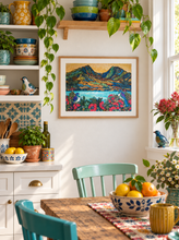 Load image into Gallery viewer, A framed landscape art print featuring colorful mountain, inlet and flowering pohutukawa with  wood pigeons displayed on a white wall in a bright, decorated kitchen or dining area with plants, bowls, and a wooden table.