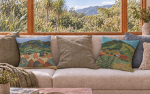 Load image into Gallery viewer, Two square throw pillows with colorful landscape prints are styled on a beige sofa in a bright room with a large window showing a lush green outdoor view.