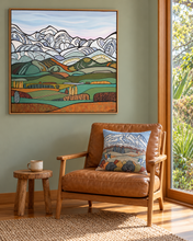 Load image into Gallery viewer, A framed landscape painting by Miranda J Caird  hangs on a sage green wall above a brown leather armchair with a matching decorative pillow. The artwork depicts stylized mountains and farmland  