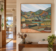Load image into Gallery viewer, A framed abstract landscape painting with layered green, blue, tan, and orange hills hangs on a beige wall above a wooden console table in a styled home interior.