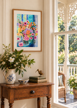 Load image into Gallery viewer, A framed colorful floral painting hangs on a cream wall above a wooden console table decorated with a blue-and-white pitcher, flowers, books, and a small bird figurine. A bright porch and garden are visible through the window.