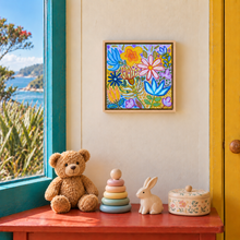 Load image into Gallery viewer, A colorful framed floral painting hangs on a cream wall above a red wooden table in a child's room. The room contains a brown teddy bear, a wooden stacking toy, a white rabbit figurine, and a floral-patterned storage box.