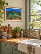 Load image into Gallery viewer, A framed landscape art print depicting green rolling hills under a blue sky, styled on a kitchen wall above a wooden countertop with a farmhouse sink, plants, and ceramic dishes.