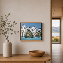Load image into Gallery viewer, A framed landscape art print depicting stylized mountains and a lake, hanging on a beige wall in a modern interior setting with a wooden bowl and a vase with branches on a table below.